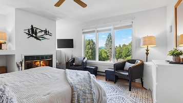 A modern bedroom with a fireplace, a large bed, a seating area with a view of the outdoors, and a television on the wall.