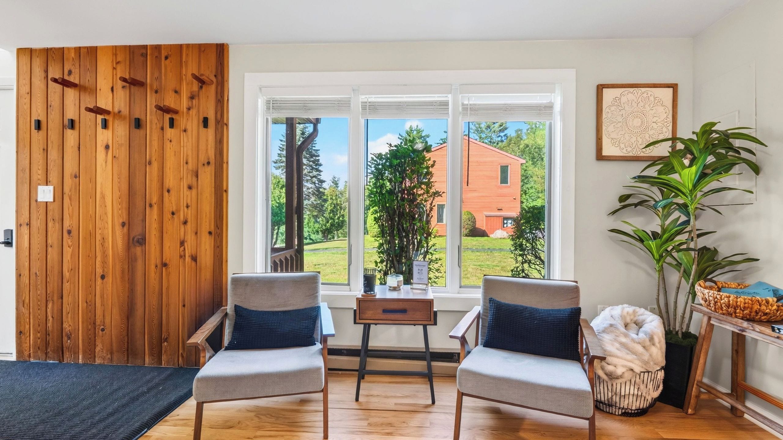 A room with two chairs, a table, a wooden door, and a window with a view of a house.