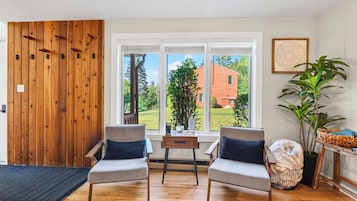 A room with two chairs, a table, a wooden door, and a window with a view of a house.