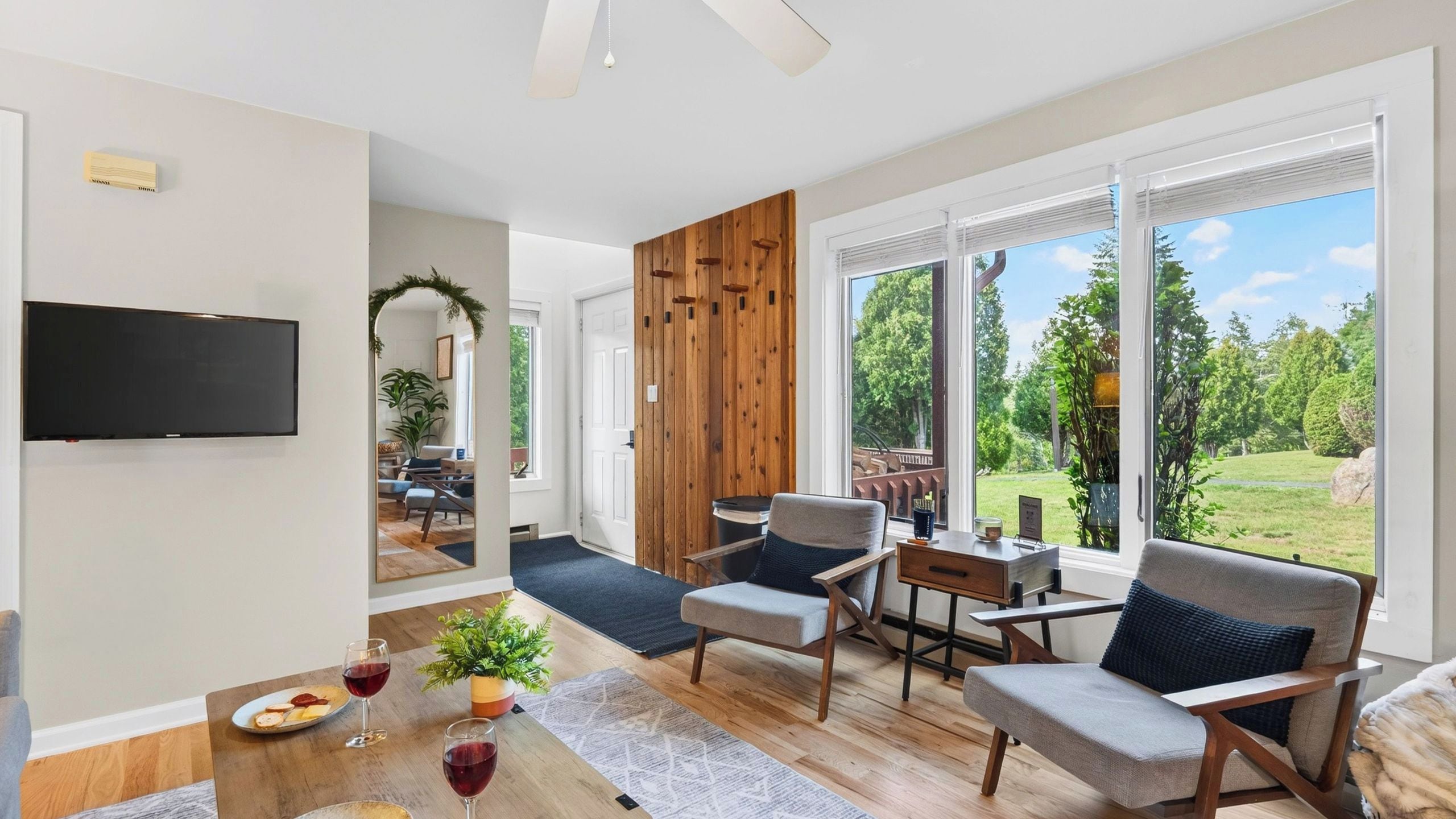 A modern living room with a flat-screen TV, comfortable seating, a dining area with a table set for two, and a large window offering a view of the outdoors.