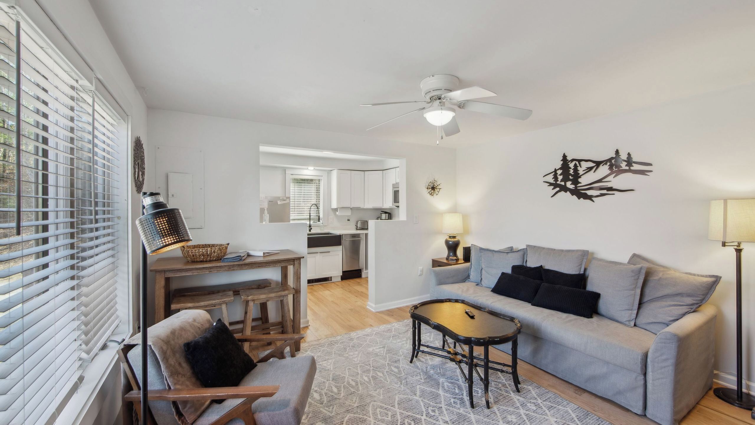 A modern living room with a sofa, a coffee table, a dining area with a table and chairs, a kitchen in the background, and a ceiling fan.