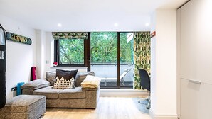 Ein modernes Wohnzimmer mit einer Couch, einem Hocker und Blick auf Grün hinter großen Fenstern.
