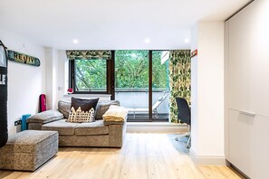 Ein modernes Wohnzimmer mit einer Couch, einem Hocker und Blick auf Grün hinter großen Fenstern.