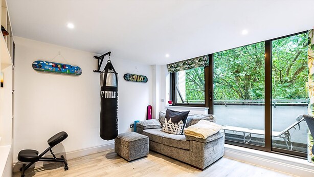 Ein modernes Wohnzimmer mit grauem Sofa, einem Faustball, einer kleinen Trainingsbank und einem großen Fenster mit Blick auf Grünflächen.