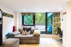 Ein modernes Wohnzimmer mit einem grauen Ecksofa, einem kleinen Hocker, einer roten Yogamatte und einem Schreibtisch mit Stuhl.