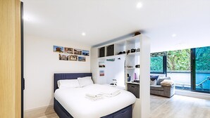 Ein modernes Schlafzimmer mit einem großen Bett, Einbauschränken, einer kleinen Sitzecke und Blick auf Grün hinter großen Fenstern.
