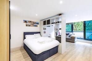 Ein modernes Schlafzimmer mit einem großen Bett, Einbauschränken, einer kleinen Sitzecke und Blick auf Grün hinter großen Fenstern.