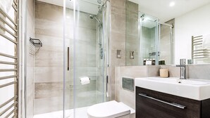 Ein modernes Badezimmer mit Duschabtrennung aus Glas, einer weißen Toilette, einem dunklen Holz-Waschtisch mit Spiegel und einem Handtuchhalter.