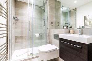 Ein modernes Badezimmer mit Duschabtrennung aus Glas, einer weißen Toilette, einem dunklen Holz-Waschtisch mit Spiegel und einem Handtuchhalter.