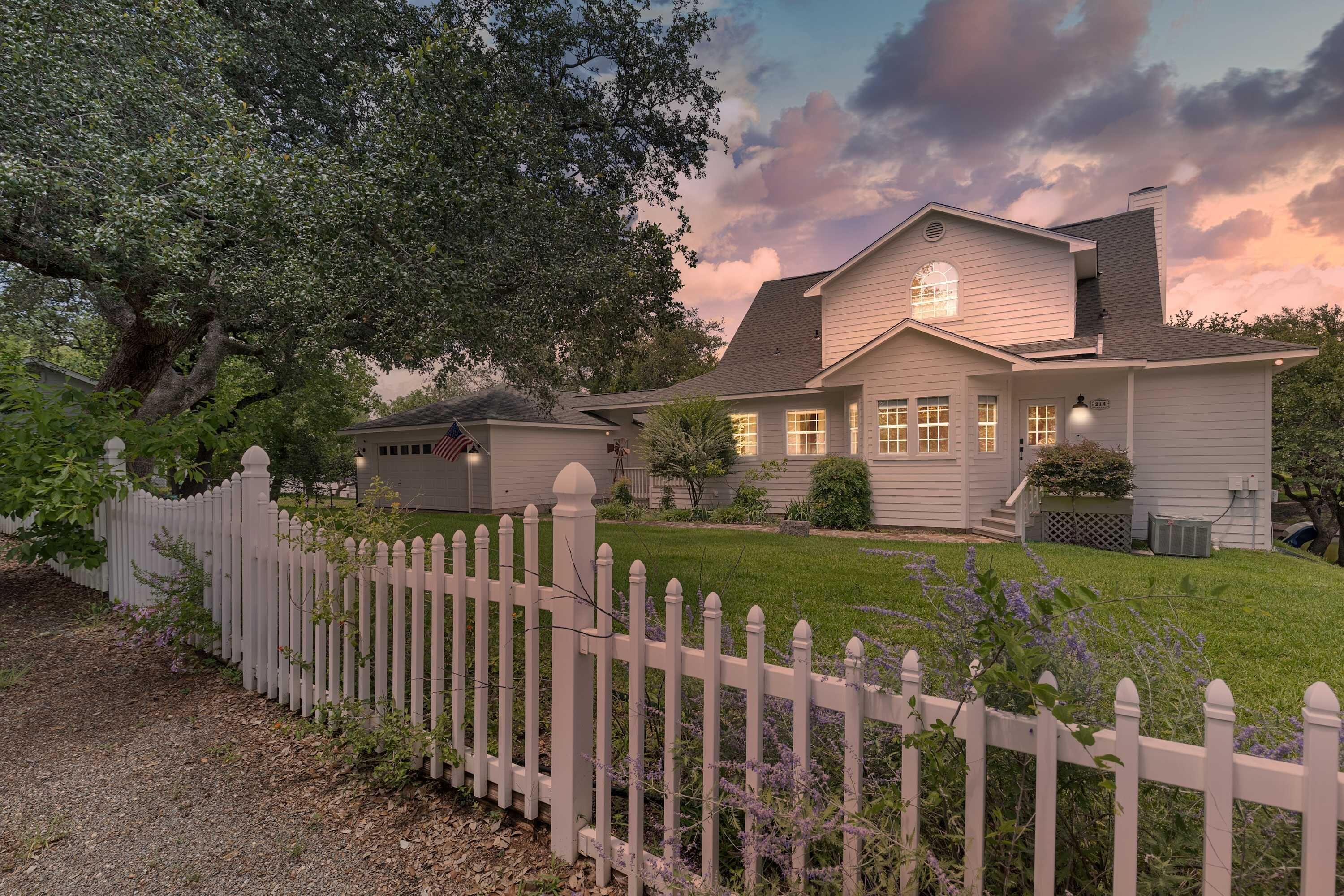 A two-story house with a white picket fence, a well-maintained lawn, and a large tree in the background.