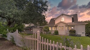 Una casa a due piani con una recinzione bianca a pali, un prato curato e un grande albero sullo sfondo.