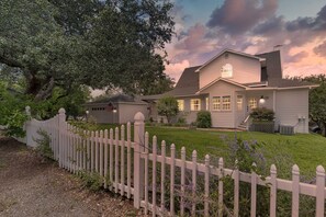 Ein zweigeschossiges Haus mit weißem Zaun, einem gepflegten Rasen und einem großen Baum im Hintergrund.