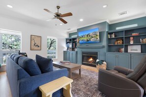 Ein Wohnzimmer mit einer Couch, einem Sessel, einem Fernseher, einem Kamin, Bücherregalen und einem Deckenventilator.