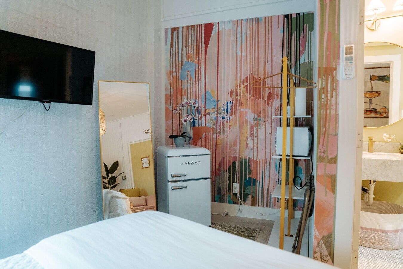 A bedroom with a bed, a dresser, a television mounted on the wall, and a bathroom with a sink and mirror.