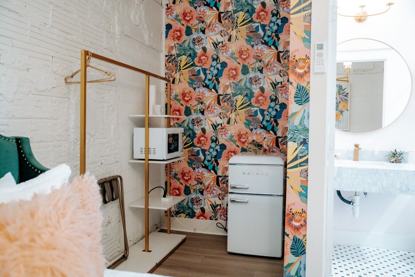 Une pièce avec un mur en briques blanches, un plancher en bois, un lit avec une tête de lit verte, un micro-ondes blanc, un réfrigérateur blanc, une étagère en bois et une salle de bain avec un lavabo et un miroir blancs.