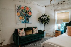Una habitación con un sofá verde, un cuadro de flores, una planta grande y una cama con cabecera blanca.