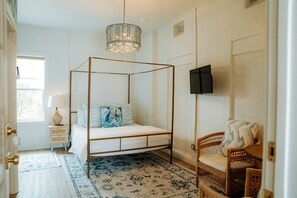 Una cama con dosel con candelabro, televisor y una silla de mimbre en una habitación con alfombras estampadas y una ventana.