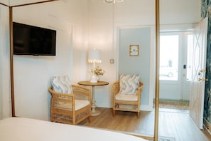 A room with two wicker chairs, a small round table with a lamp, a television mounted on the wall, and a door leading outside. - Room #5 The Oleander Hotel Galveston (Galveston)