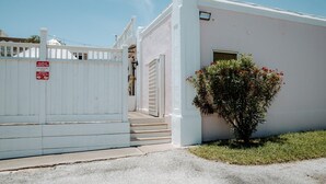 Un edificio con fachada blanca, entrada protegida por una reja y un arbusto con flores rojas.