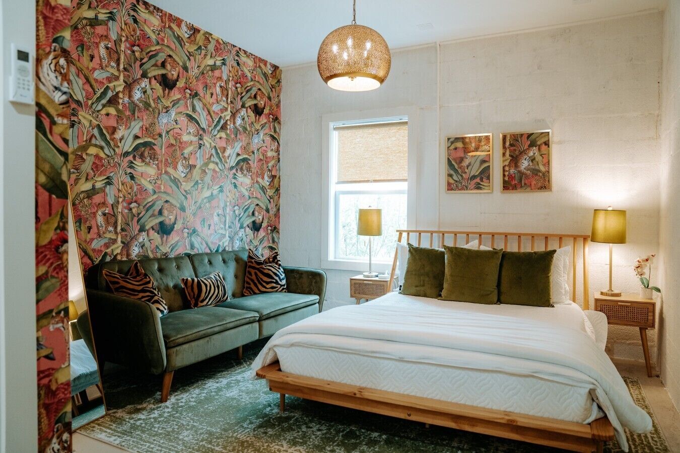 Un dormitorio con una cama, un sofá verde, un papel tapiz estampado, una ventana y dos lámparas de noche.