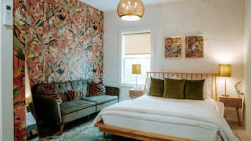 Un dormitorio con una cama, un sofá verde, un papel tapiz estampado, una ventana y dos lámparas de noche.