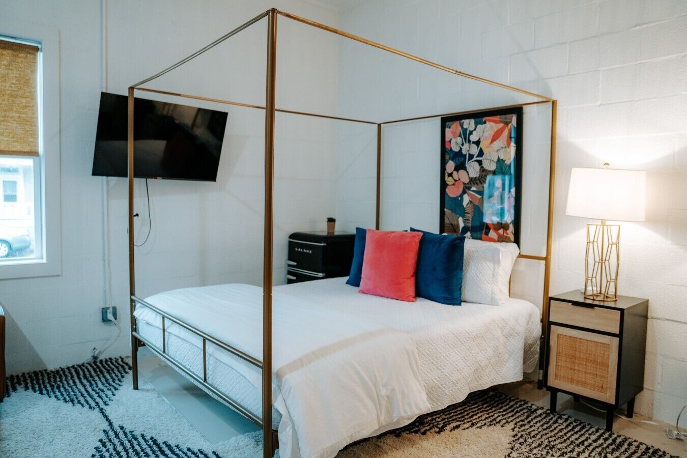 A four-poster bed with a television above it, a nightstand with a lamp, and a floral painting on the wall.