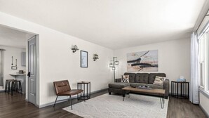 A modern living room with a sofa, a chair, a coffee table, and a rug. There is a painting on the wall and a small table with a lamp.