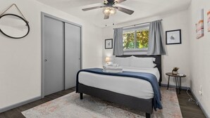 A bedroom with a bed, a ceiling fan, a window with curtains, a small table with a plant, and a door.