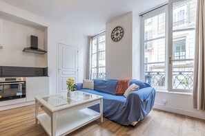 Ein modernes Wohnzimmer mit einer Couch, einem Couchtisch, einer Küchenzeile mit Backofen und großen Fenstern mit Blick auf Gebäude.
