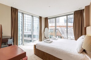 A hotel room with a large bed, a desk, a chair, a mirror, and a view of buildings through large windows. - Aldgate East Apartment With Balcony (London)