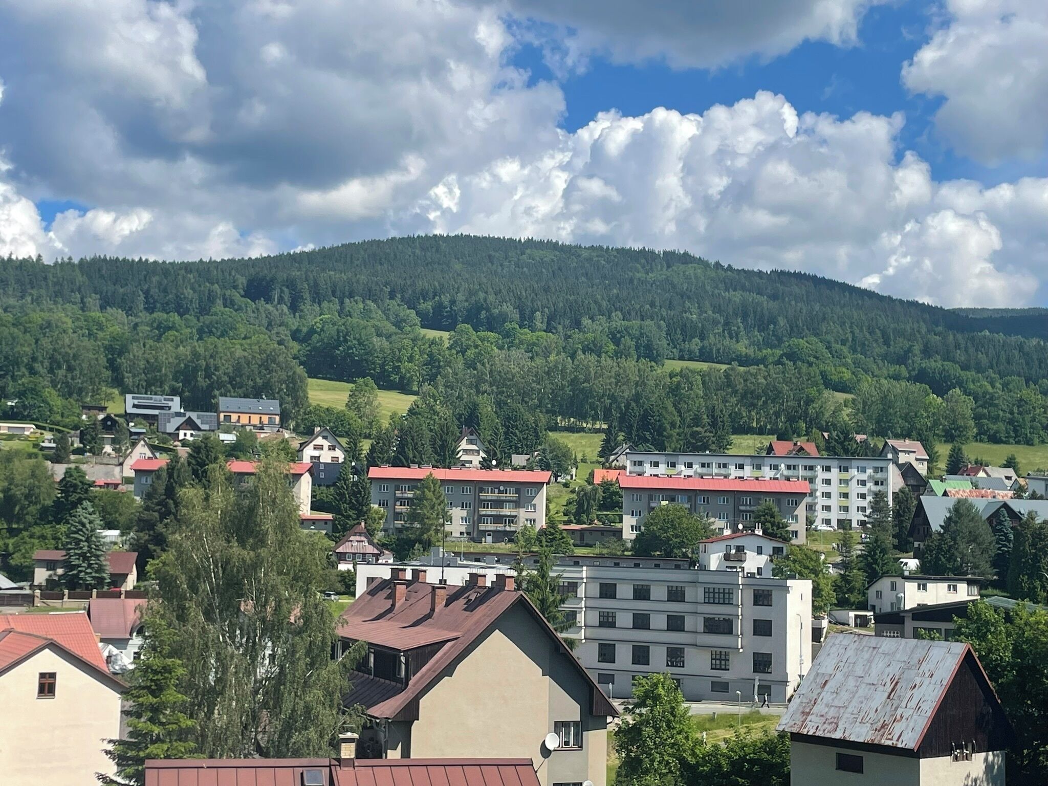 A town with buildings, including a large multi-story building with a red roof, surrounded by greenery and hills.