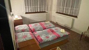 Um quarto com cama, duas janelas com cortinas, um radiador, uma cadeira e uma mesa de cabeceira com abajur.