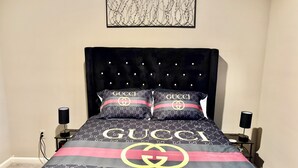 Uma cama com cabeceira preta, duas almofadas com o logo da Gucci e um cobreleito com o logo da Gucci.