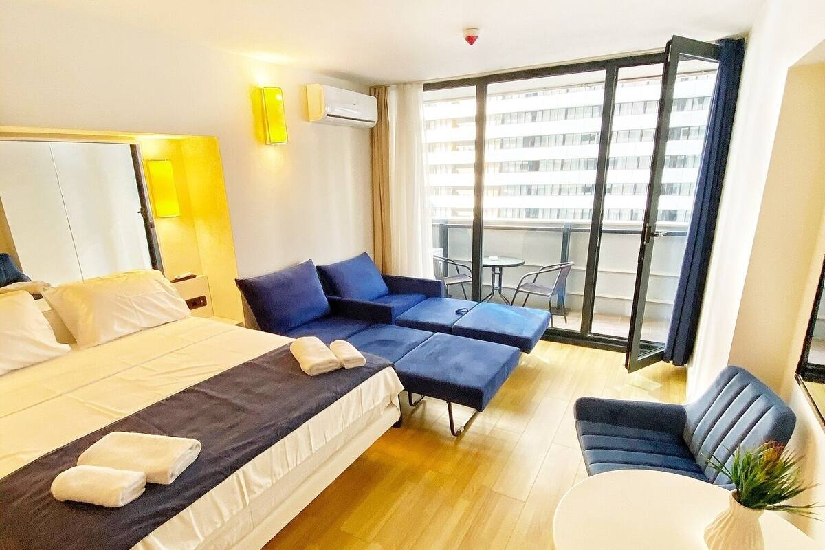 Habitación de hotel con una cama grande, dos sofás azules, una mesita blanca y un balcón con vistas a edificios.