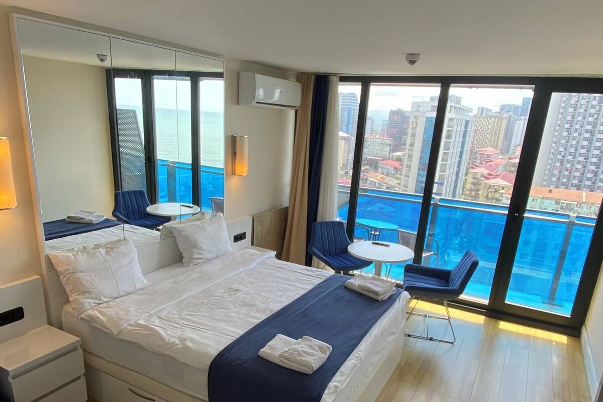 Una habitación de hotel moderna con una cama grande, una mesita, una silla y un balcón con vistas a la ciudad.