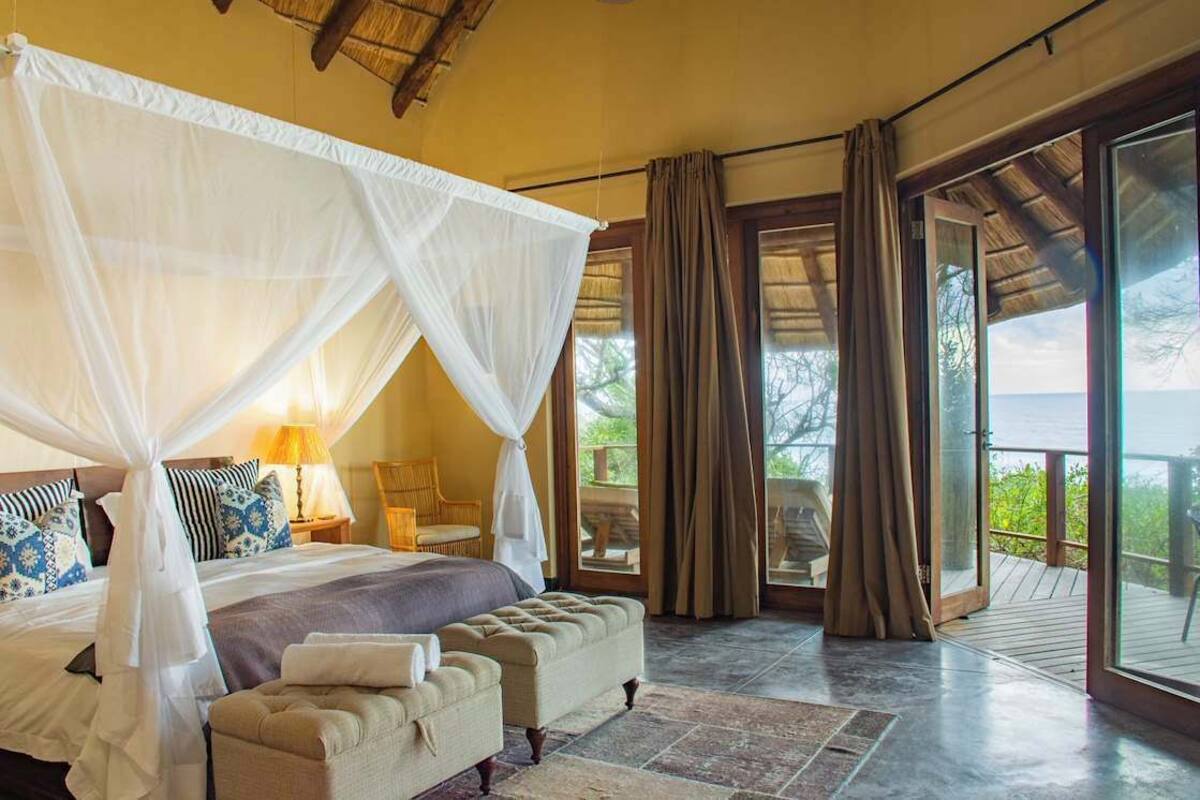 A bedroom with a four-poster bed, a sitting area with a sofa and armchairs, and a balcony with a view.