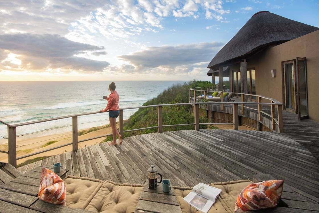 A wooden deck with a view of the ocean, a thatched-roof building, and a person standing by the railing.