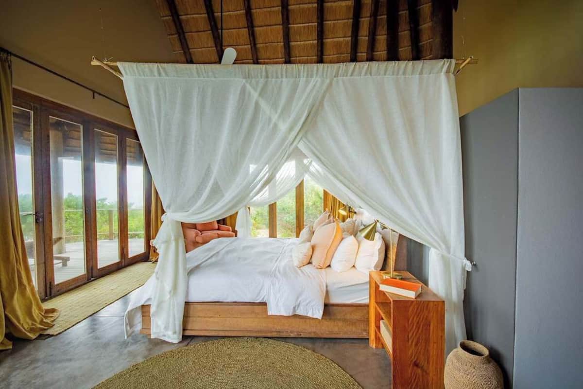 A bedroom with a canopy bed, a wooden headboard, a bedside table with a book, and a view of greenery through large windows.