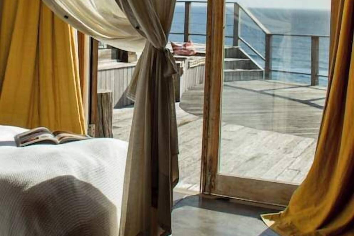A room with a bed, a window with curtains, and a view of a balcony and ocean.