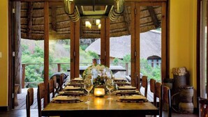 Una mesa de comedor lista para una comida, con sillas, una araña y vista a una construcción de paja en el exterior.