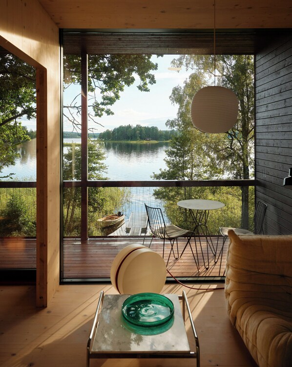 A room with a large window overlooking a lake, a round table, and a sofa. - Architect-designed cabin with Harefjord views (Säffle)