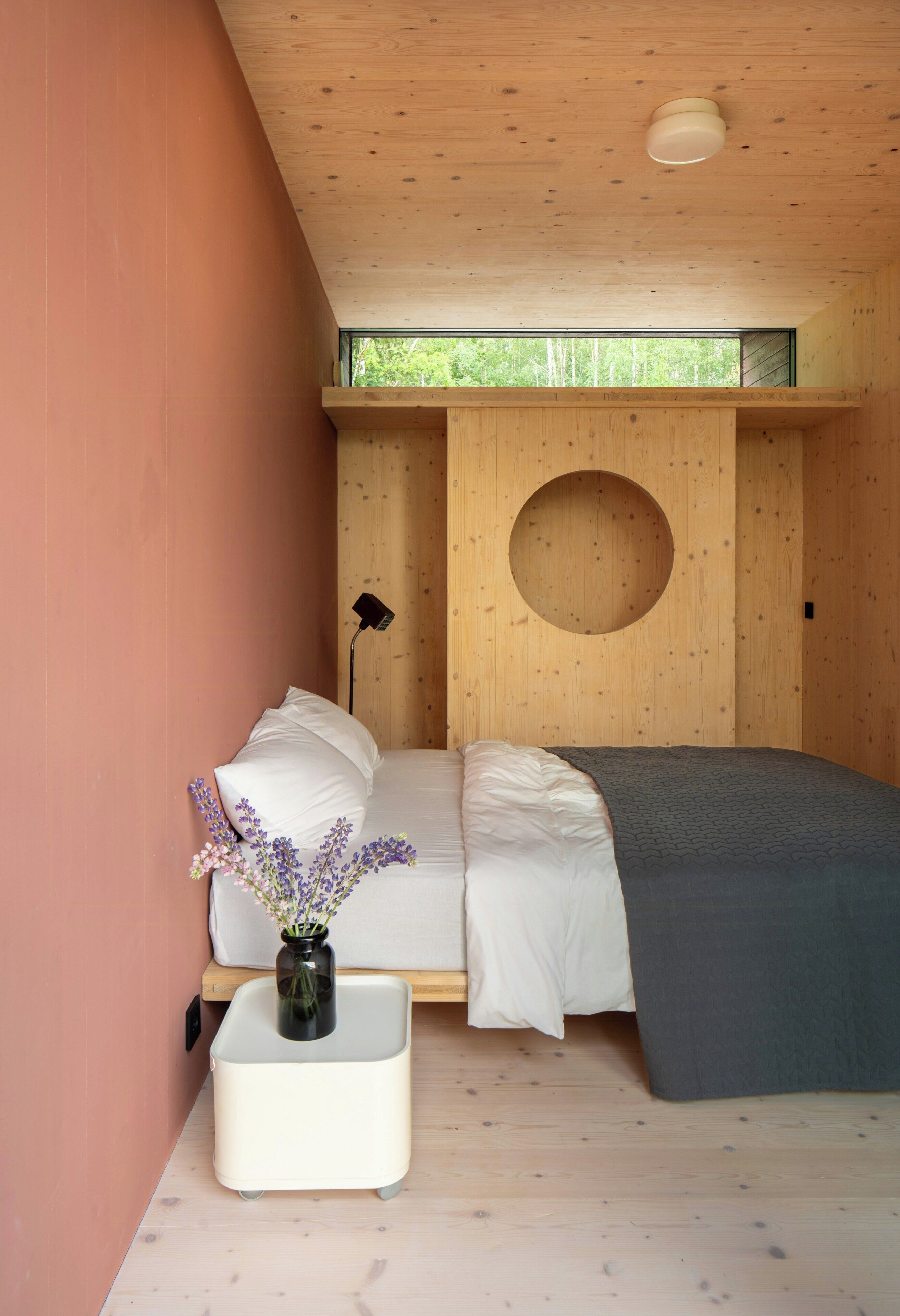 A bedroom with a bed, a bedside table with a vase, and a circular cutout on the wall.