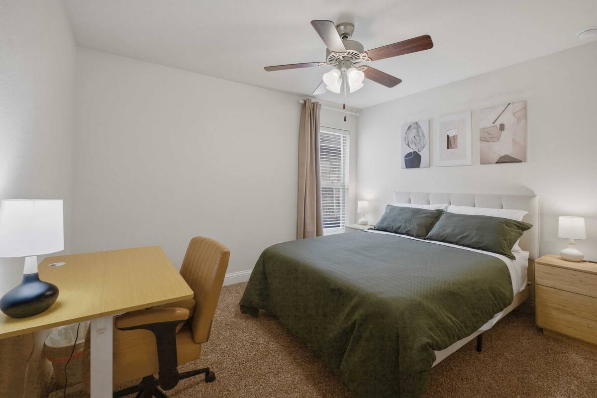 Un dormitorio con cama, escritorio, silla, mesita de noche, ventilador de techo y ventana con persianas.