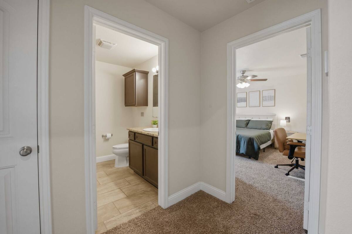 Un pasillo que conduce a un baño con inodoro, lavabo y armario, y un dormitorio con cama, silla y escritorio.