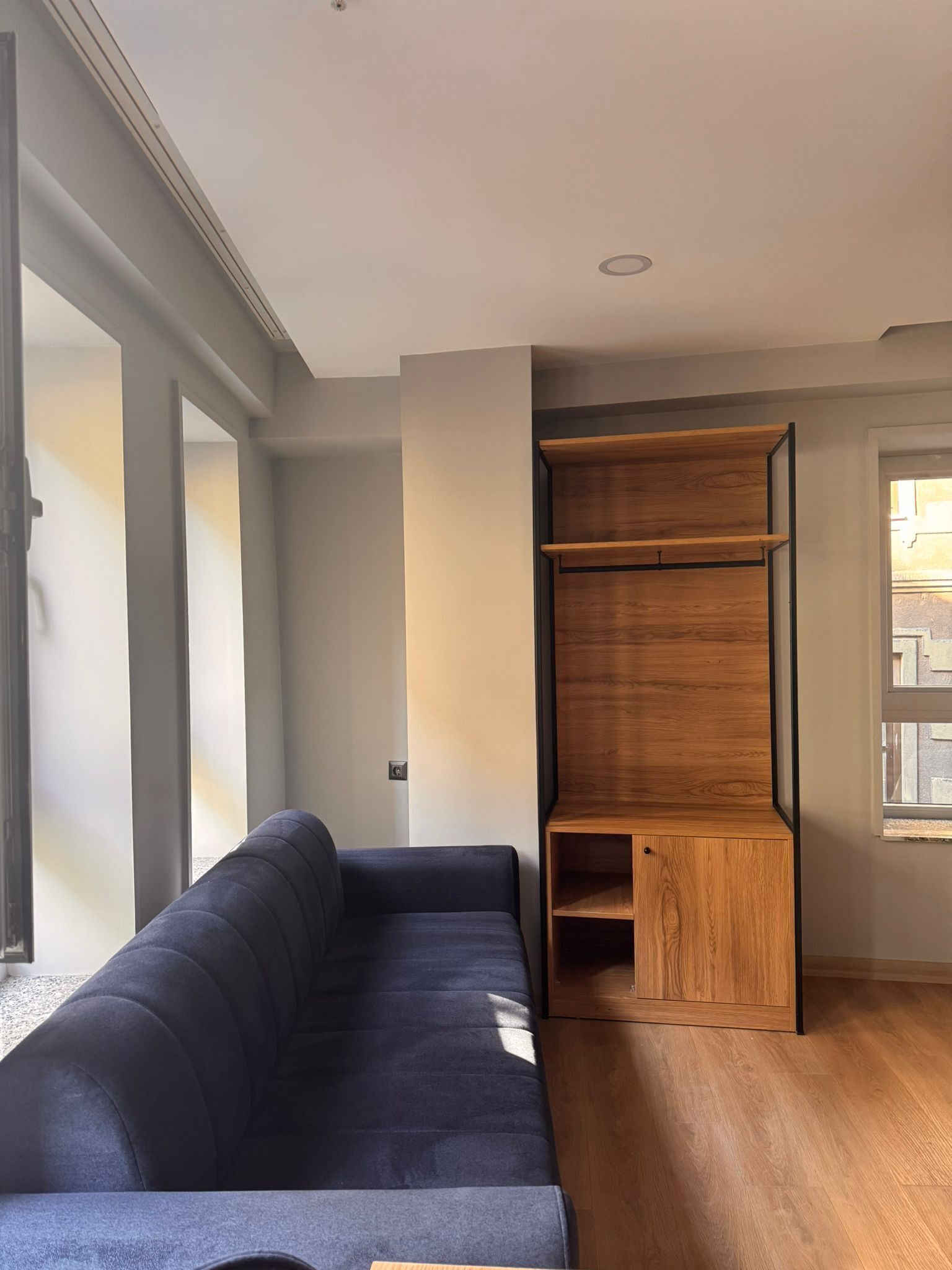A modern room with a dark blue sofa, wooden shelving unit, and large windows.