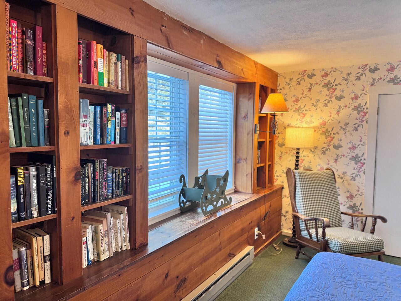 Ein Zimmer mit Holzregal, Jalousien, einem Sessel und einer Lampe.