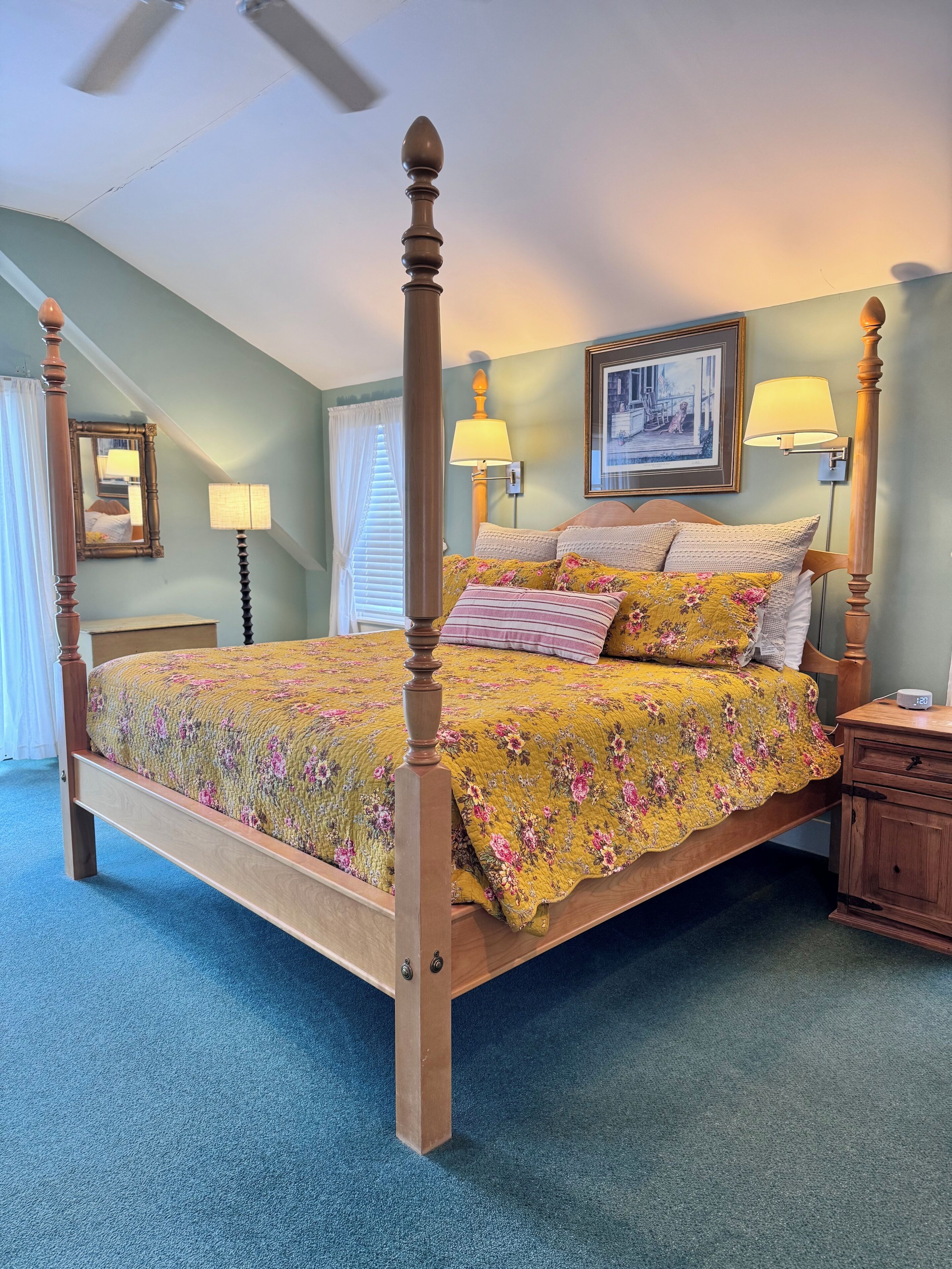 A four-poster bed with a floral bedspread, two bedside lamps, a nightstand, a framed picture on the wall, and a ceiling fan.