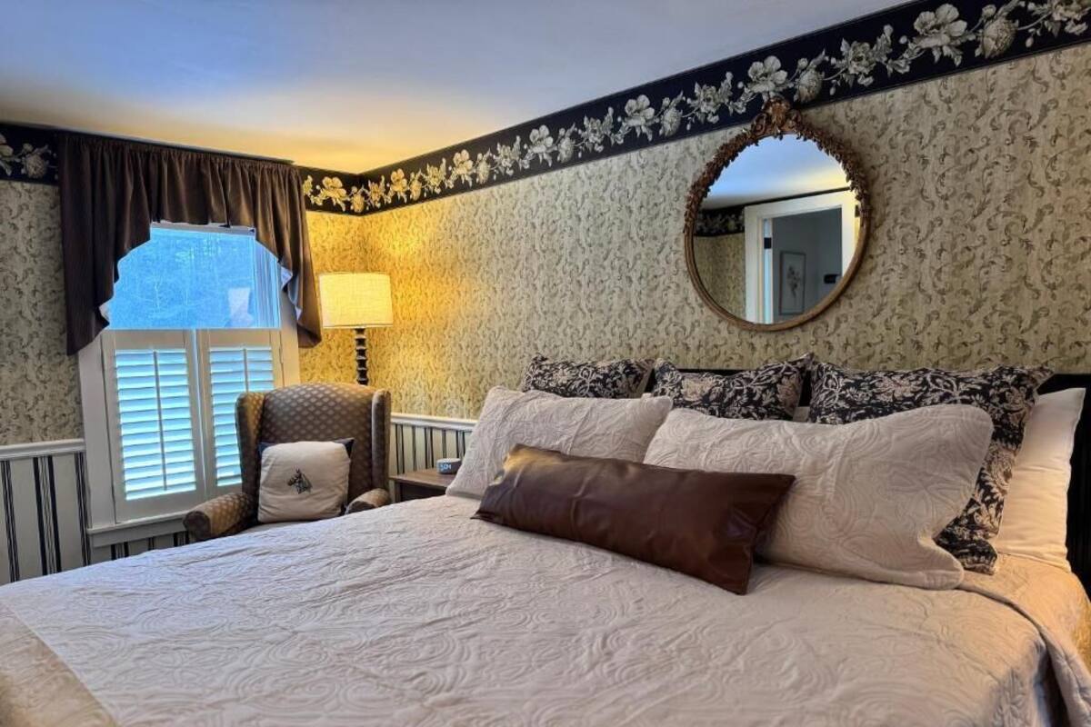 A bedroom with a large bed, a round mirror, a window with curtains, and a wall with floral wallpaper.