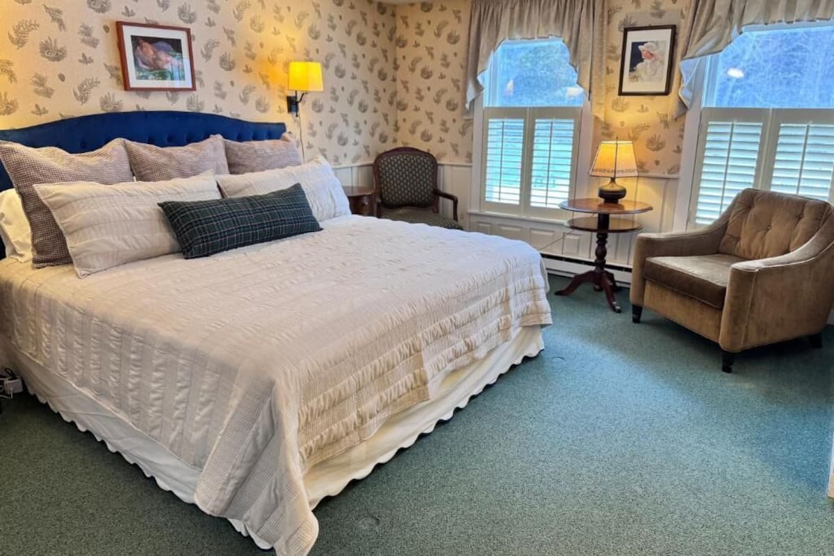 A bedroom with a large bed, a chair, a small table, and two windows with curtains.