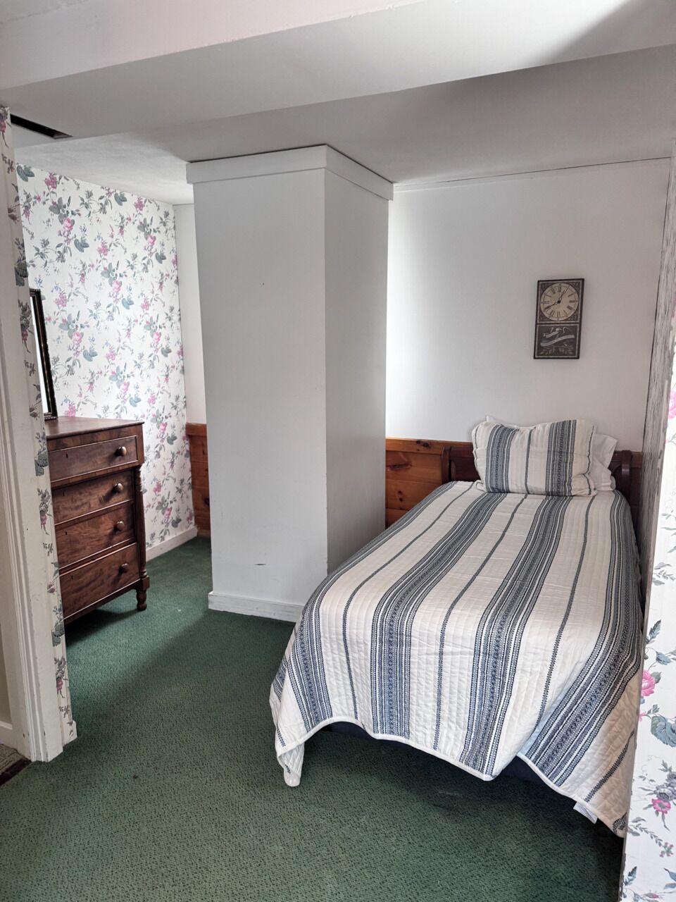 A bedroom with a bed, a dresser, a floral wallpapered wall, and a clock on the wall.